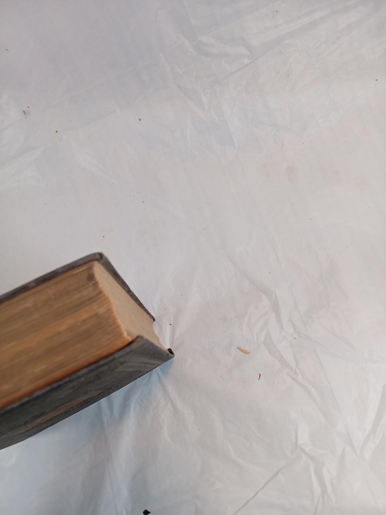 May include: A partially visible, aged book with a dark blue cover and aged, tan-colored pages. The book's edges show wear, suggesting it is an antique or vintage item. The book is positioned at an angle against a white, textured background.