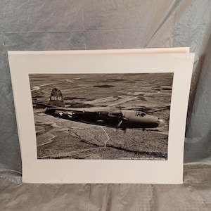 May include: Black and white photograph of a vintage airplane in flight over a landscape. The aircraft has two propellers and the number "015 42" is visible on the tail. The image is framed in a white border.