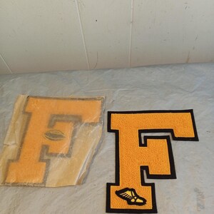 May include: Two yellow felt letter "F" patches with black borders. One patch has a football graphic, the other has a shoe graphic. The patches are likely for a varsity jacket or other apparel.