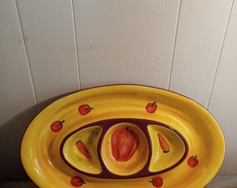 Puede incluir: Plato ovalado para servir con exterior amarillo y tres secciones interiores. El plato presenta un diseño pintado de pimientos rojos y pequeñas manzanas rojas. El plato está diseñado para servir aperitivos o snacks.