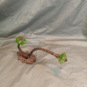 May include: A wall-mounted hook featuring a bronze-toned metal base with an ornate, leaf-like design. Two twisted metal arms extend from the base, each topped with a faceted, translucent lime green glass knob in a flower and diamond shape.