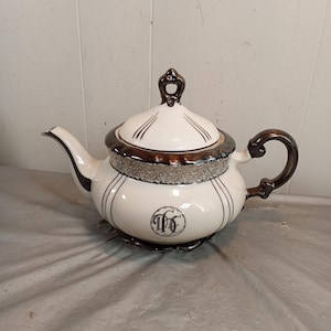 May include: Cream-coloured teapot with dark brown handle, spout, and lid accents. The lid features a decorative finial and vertical lines. The body has black vertical lines and a monogram. The teapot sits on a silver-coloured base.