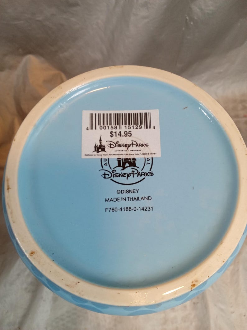 Pu&ograve; includere: Base di una tazza Disney Parks in ceramica azzurra con bordo bianco. Il fondo della tazza presenta il logo Disney Parks, la scritta "&copy;DISNEY MADE IN THAILAND" e un codice a barre.