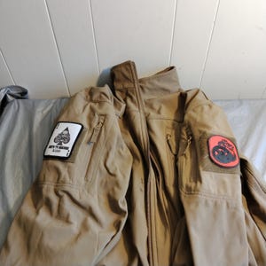 May include: Tan tactical jacket with multiple pockets and patches. One patch features a skull and ace of spades, the other a red and black design. The jacket has a high collar and a full-length zipper.