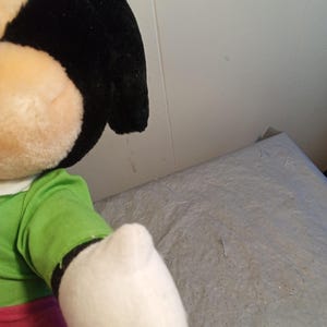May include: A plush doll featuring a black head, large ears, and a tan face. The doll wears a green shirt, red pants, and a white glove. The doll is a cartoon character.