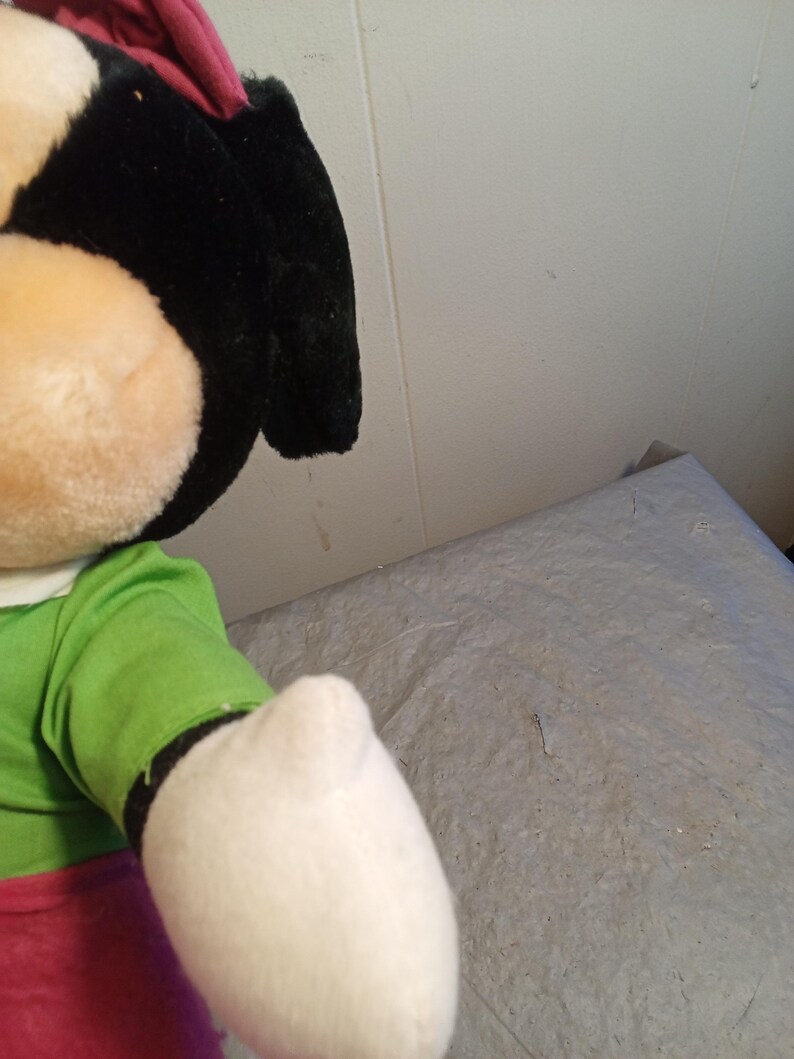 May include: A plush doll featuring a black head, large ears, and a tan face. The doll wears a green shirt, red pants, and a white glove. The doll is a cartoon character.