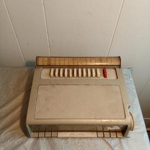 Op de afbeelding: Een vintage, crèmekleurige speelgoedpiano met een reeks witte toetsen en een rode knop. De piano heeft een luidsprekergedeelte en een reeks kleine toetsen aan de onderkant. Het bovenste gedeelte heeft een reeks kleine, rechthoekige toetsen. Het speelgoed is van plastic.