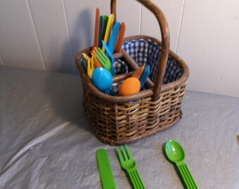 May include: A wicker basket with a handle, lined with blue and white checkered fabric, filled with colorful plastic utensils.  Three green plastic utensils, a knife, fork, and spoon, are laid out on a white surface.