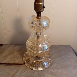 May include: A clear glass table lamp with a decorative, layered design. The lamp features a brass-colored bulb socket and a brown electrical cord. The lamp's base and body are made of clear, molded glass.