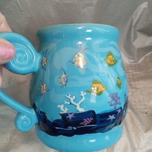 May include: A light blue ceramic mug with a whimsical underwater scene. The mug features illustrations of colorful fish, coral, and seaweed. The handle is a matching light blue with a decorative swirl.