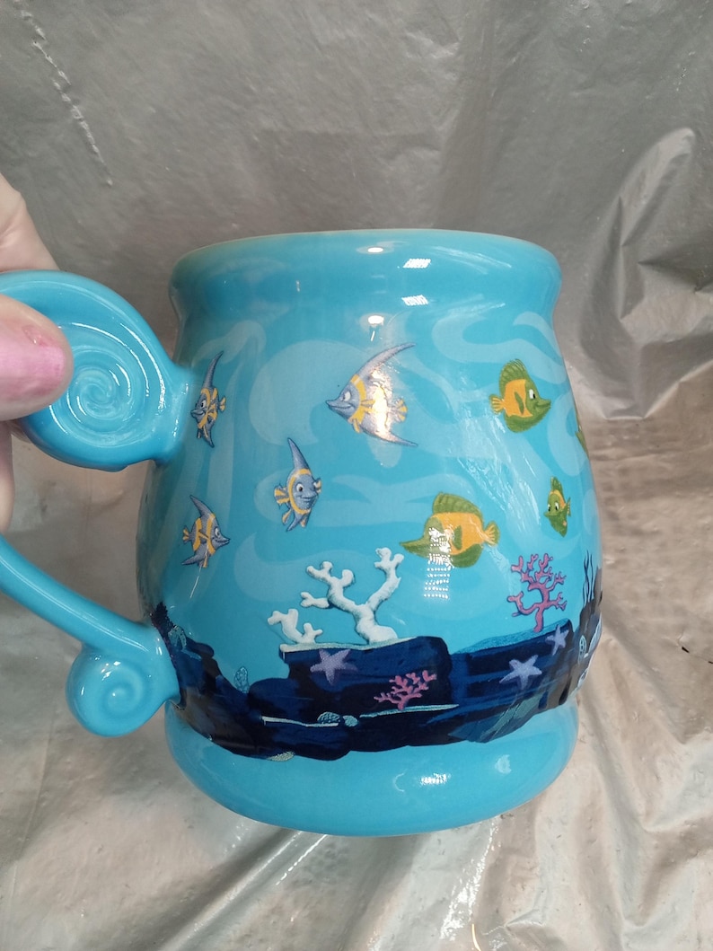 May include: A light blue ceramic mug with a whimsical underwater scene. The mug features illustrations of colorful fish, coral, and seaweed. The handle is a matching light blue with a decorative swirl.