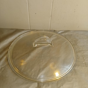 Op de afbeelding: Een helder glazen deksel met een handvat. Het ronde deksel heeft een zilveren rand en is ontworpen voor een pot of pan. De handgreep is een klein, gebogen stuk glas.