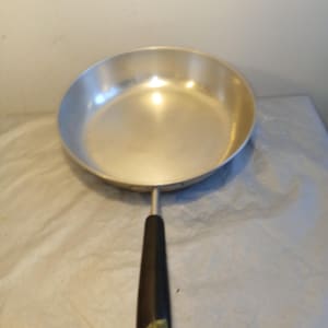 May include: A silver-colored frying pan with a black handle. The pan has a rounded shape and a long handle. The handle is black and has a metal ring at the end. The pan is made of metal and is designed for cooking.