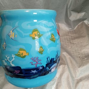 May include: A light blue ceramic mug with an underwater scene featuring yellow and green fish, coral, and seaweed. The mug has a wide, rounded shape and a curved handle. The design is a cartoon style.