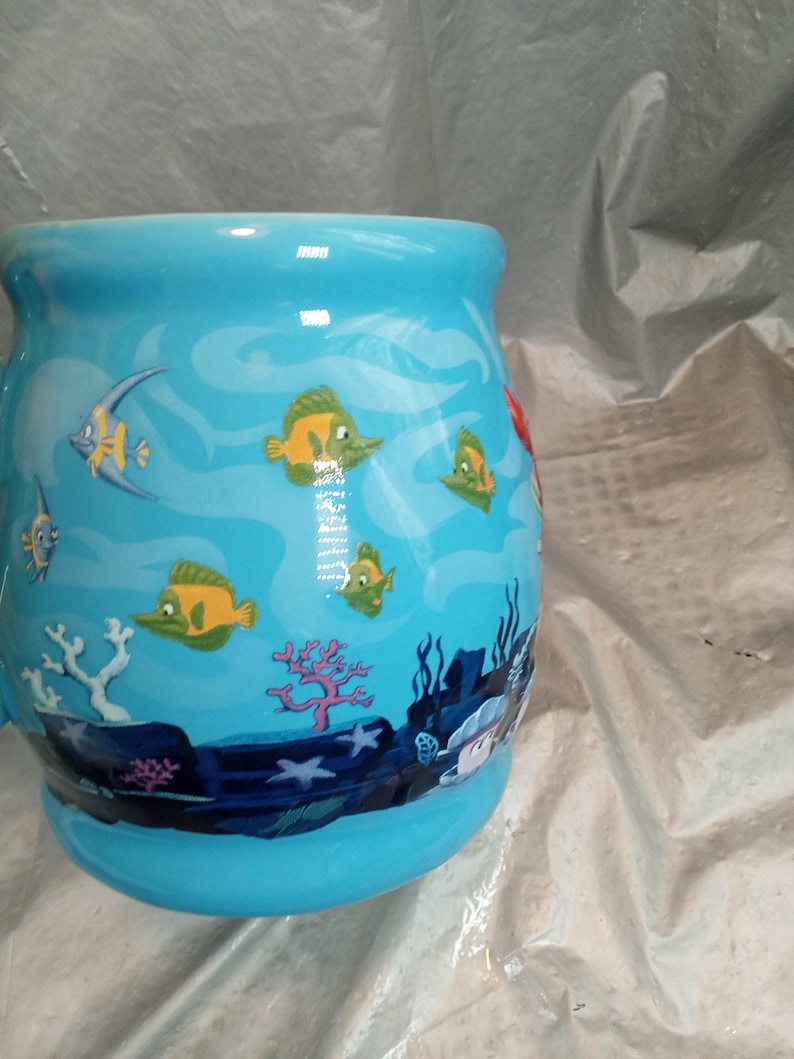May include: A light blue ceramic mug with an underwater scene featuring yellow and green fish, coral, and seaweed. The mug has a wide, rounded shape and a curved handle. The design is a cartoon style.
