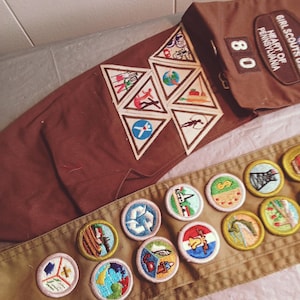 May include: A brown Girl Scout sash and uniform pieces with various embroidered patches. The sash displays several round and triangular patches with colorful designs. The uniform includes a pocket with the text "Girl Scouts of Heart of Pennsylvania" and the number "80".
