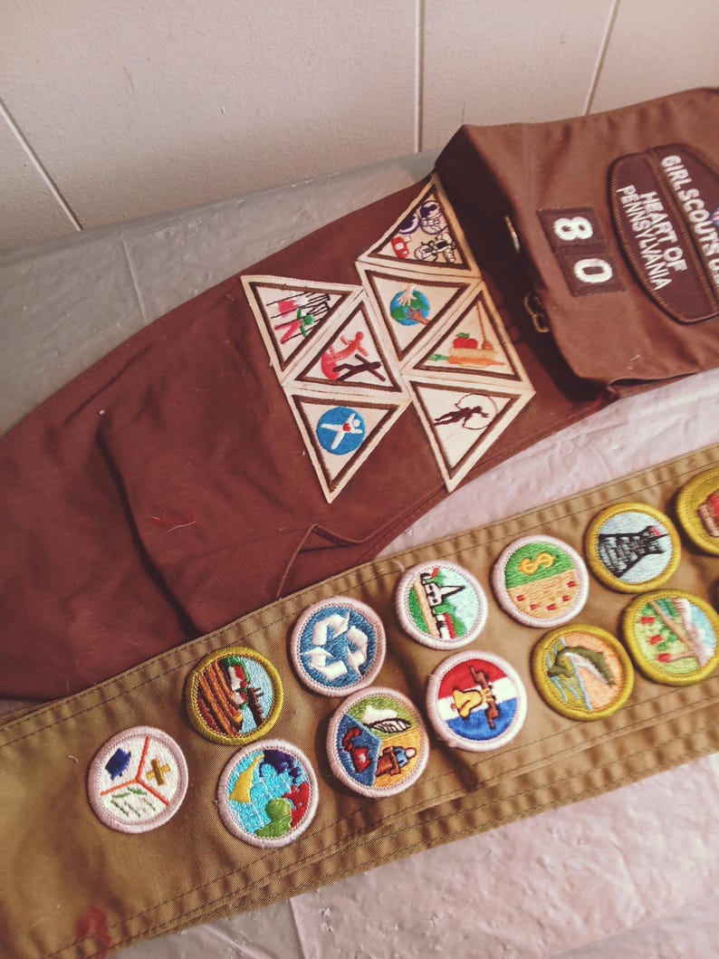 May include: A brown Girl Scout sash and uniform pieces with various embroidered patches. The sash displays several round and triangular patches with colorful designs. The uniform includes a pocket with the text "Girl Scouts of Heart of Pennsylvania" and the number "80".