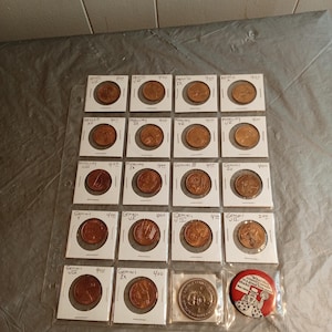 May include: A collection of vintage coins and a button. The coins are in protective sleeves, labeled with names like "Apollo" and "Gemini." The button is red with a cartoon image and text.