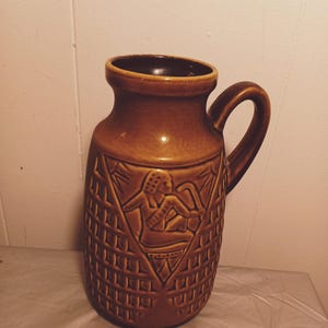 May include: A brown ceramic vase with a geometric pattern and a raised design of a person in a triangular shape. The vase has a wide mouth and a handle.