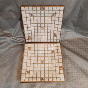 Puede incluir: Dos bandejas decorativas cuadradas con un diseño de mosaico de azulejos blancos y ribete dorado. Los azulejos están dispuestos en un patrón de cuadrícula, con azulejos de acento dorados. Las bandejas están abiertas e inclinadas, mostrando el diseño interior.