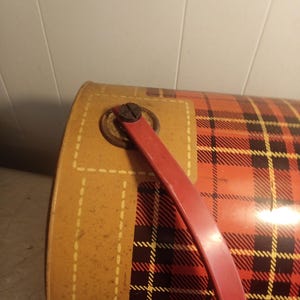 May include: Vintage metal container with a red and black plaid pattern. The container has a red handle and a tan-colored side with a stitched design. The container is likely used for storage or carrying items.