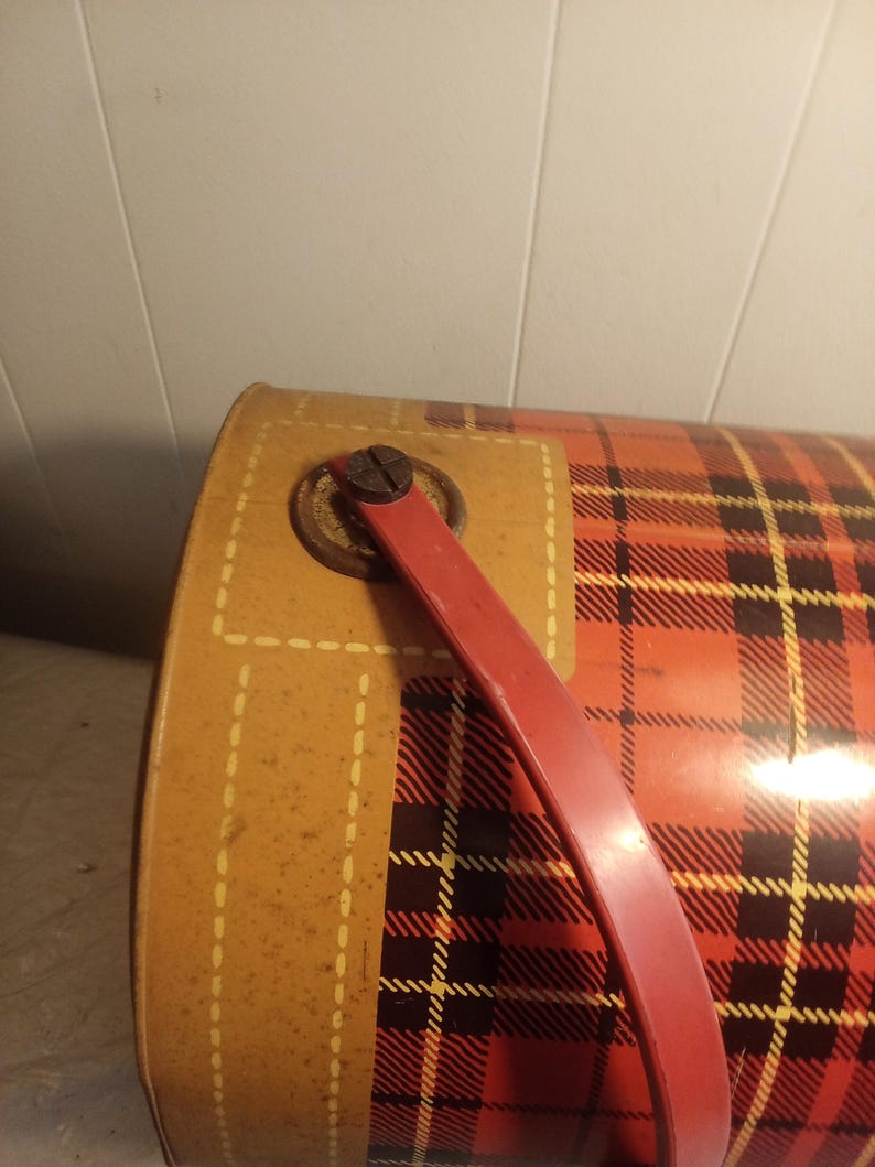 May include: Vintage metal container with a red and black plaid pattern. The container has a red handle and a tan-colored side with a stitched design. The container is likely used for storage or carrying items.
