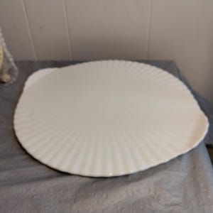 Pode incluir: Uma travessa grande, de cor branco-sujo, com borda ondulada. A travessa tem um design canelado em forma de leque que irradia do centro. A travessa está sobre uma superfície cinza.