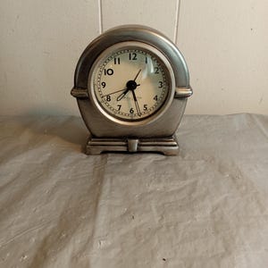 May include: A silver-coloured, vintage-style clock with a round face and a curved top. The clock face has black numerals and hands, with the brand name "Bulova" visible. The clock sits on a rectangular base.