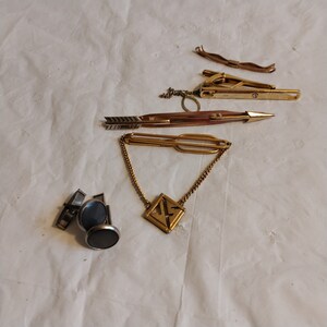 May include: A set of gold-toned tie clips and cufflinks. The tie clips feature a variety of designs, including a simple arrow, a geometric pattern, and a chain with a square pendant. The cufflinks are silver-toned and have a black stone inlay.