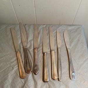 Può includere: Un assortimento di coltelli d'argento vintage con vari design del manico. Le lame sono argento opaco e i manici presentano motivi e forme ornate. I coltelli sono disposti su una superficie grigio chiaro.