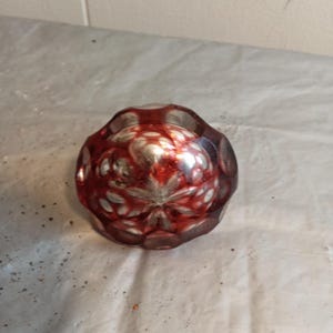 May include: A round, red glass object with a starburst design. The object has a faceted, textured surface and is likely a decorative item or paperweight. The glass has a metallic, reflective quality.