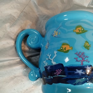 May include: A light blue ceramic mug with a whimsical underwater scene. The mug features a curved handle and is decorated with cartoon fish, coral, and aquatic plants. The design is in shades of blue, green, and yellow.