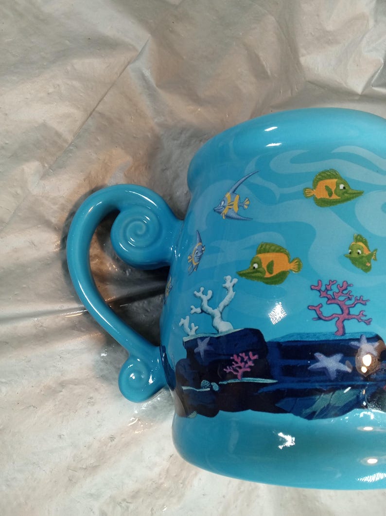 May include: A light blue ceramic mug with a whimsical underwater scene. The mug features a curved handle and is decorated with cartoon fish, coral, and aquatic plants. The design is in shades of blue, green, and yellow.