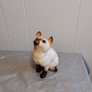 May include: A white ceramic figurine of a Siamese cat sitting with its head tilted up. The cat has brown ears, a brown nose, and blue eyes.