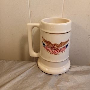 Könnte beinhalten: Cremefarbener Bierkrug aus Keramik mit Henkel und breitem Fuß. Der Krug zeigt ein patriotisches Design mit einem ausgebreiteten Adler, amerikanischen Flaggen und Text. Der Rand hat einen braunen Akzent.