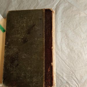 May include: An antique book with a worn, dark green cover and a brown spine. The cover shows signs of age with visible wear and tear. The spine is a darker brown, and the pages have a yellowed appearance, suggesting the book's age.