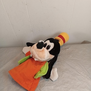 May include: A plush toy of Goofy, a Disney cartoon character, is lying on a surface. The toy has a black head, white muzzle, and a yellow hat with a red stripe. It wears an orange shirt with green sleeves.