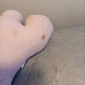 May include: A light pink plush toy with a heart-shaped design. The plush toy has two brown spots on its surface. The toy is made of a soft, fuzzy material.