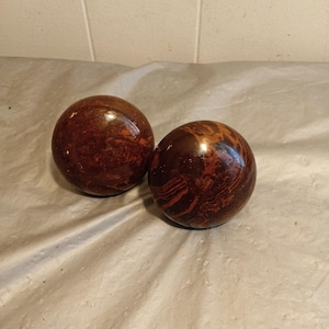 Op de afbeelding: Twee ronde, gepolijste houten bollen met een rijke, roodbruine kleur en ingewikkelde nerfpatronen. De bollen hebben een glanzende afwerking die het licht reflecteert. Ze lijken decoratief of functioneel te zijn.