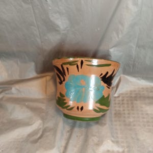 Op de afbeelding: Een kleine, handbeschilderde keramische pot met een beige basis, met een groot turquoise bloemmotief. De rand en de basis zijn groen geschilderd, met zwarte bladaccenten. De pot wordt waarschijnlijk gebruikt voor decoratieve doeleinden.