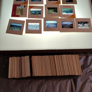 Puede incluir: Una gran pila de monturas de diapositivas de cartón vintage, algunas con notas escritas a mano, se muestran en una mesa de luz. Las diapositivas están dispuestas en filas y columnas, con algunas diapositivas individuales en la superficie de la mesa.