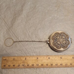 Op de afbeelding: Een zilverkleurig, rond medaillon met een bloemen filigraan ontwerp op het deksel. Het medaillon is bevestigd aan een ketting en een ring. Een houten liniaal met inch-metingen ligt naast het medaillon.
