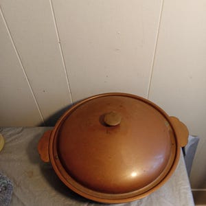 May include: A copper-colored, round cooking pot with a lid. The lid has a small, round handle and two side handles. The pot is sitting on a gray surface.