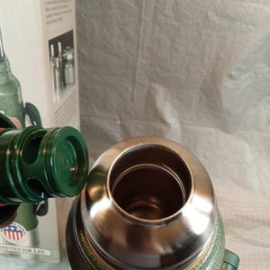May include: A green vacuum-insulated thermos with a metallic rim and a detached green plastic stopper. The thermos is positioned in front of its original cardboard packaging, which features the text America's #1 Brand and Guaranteed for Life.