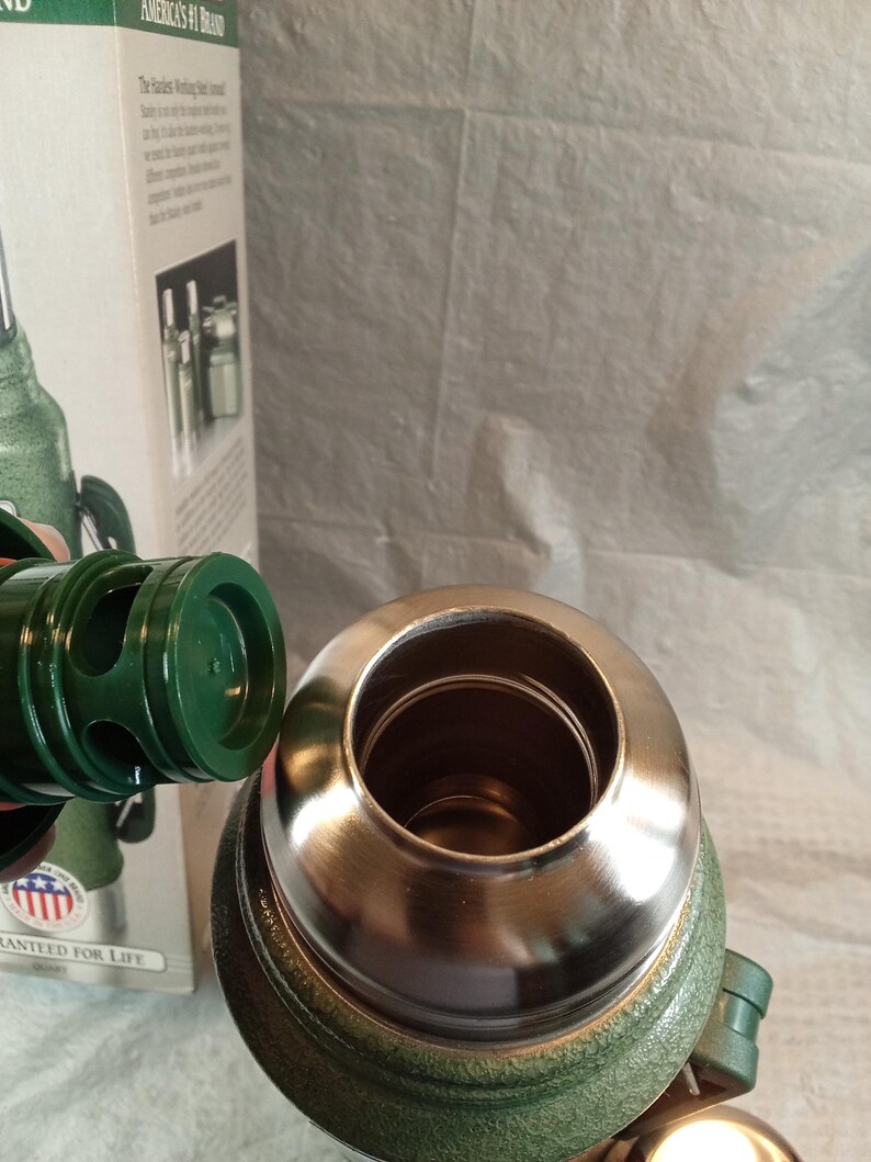 May include: A green vacuum-insulated thermos with a metallic rim and a detached green plastic stopper. The thermos is positioned in front of its original cardboard packaging, which features the text America's #1 Brand and Guaranteed for Life.