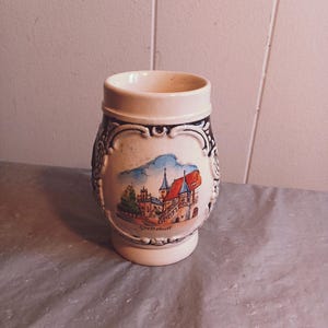 Puede incluir: Una jarra de cerveza de cerámica blanca con un diseño negro y azul. La jarra presenta una imagen pintada de un castillo con la palabra "Gottskal" escrita debajo.