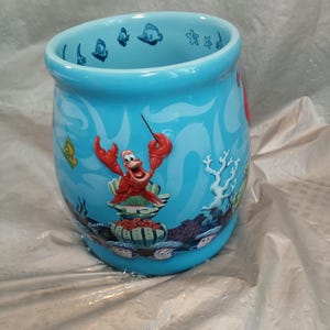 May include: A turquoise ceramic mug featuring a cartoon illustration of Sebastian the crab from The Little Mermaid. The mug has a curved shape and is decorated with underwater scenes, including fish and coral.