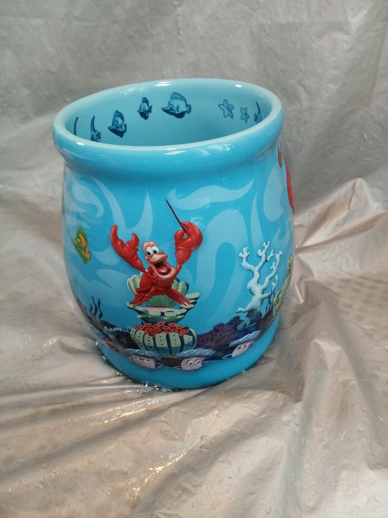 May include: A turquoise ceramic mug featuring a cartoon illustration of Sebastian the crab from The Little Mermaid. The mug has a curved shape and is decorated with underwater scenes, including fish and coral.