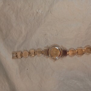 May include: A gold-tone wristwatch featuring a circular case and a link bracelet composed of alternating circular and square metal segments. The watch back is plain and metallic, and the bracelet includes a standard clasp mechanism for closure.