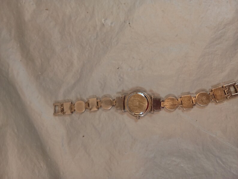 May include: A gold-tone wristwatch featuring a circular case and a link bracelet composed of alternating circular and square metal segments. The watch back is plain and metallic, and the bracelet includes a standard clasp mechanism for closure.